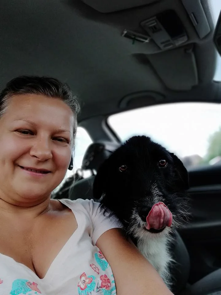 Mirela smiling in her car while a black dog licks its nose over her shoulder.
