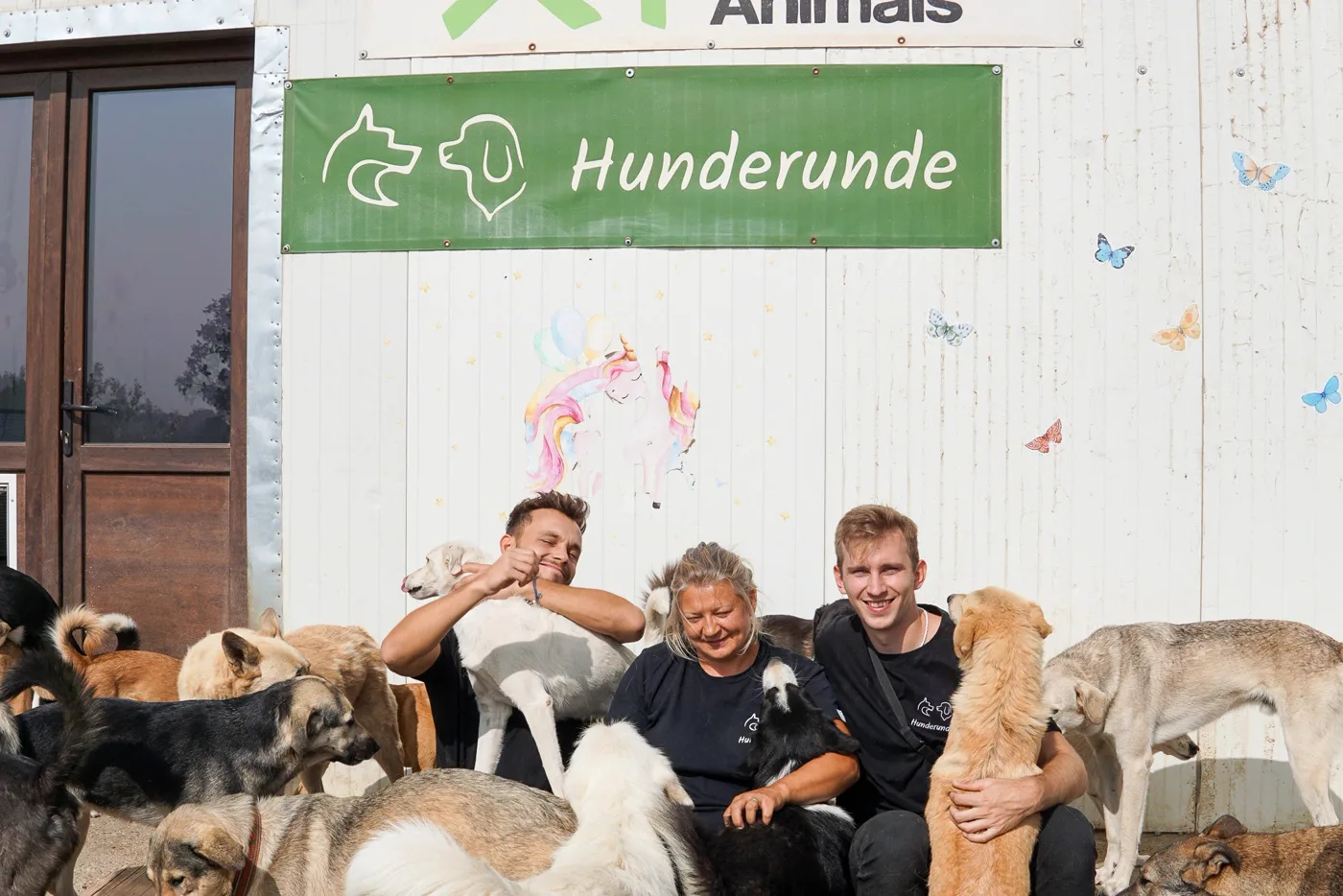 Fabio, Luis and Mirela sitting together with the dogs in front of the lodge, a Hunderunde banner on the wall.